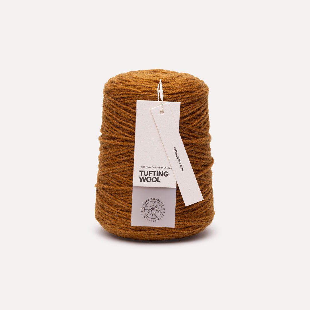 A cone of mustard brown tufting wool yarn with two white paper tags attached, set against a clean, light off-white background.