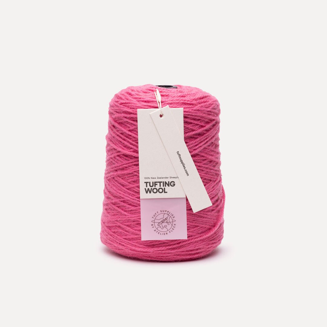 A cone of bright pink tufting wool yarn with a white paper hangtag, photographed on a clean, light off-white background.