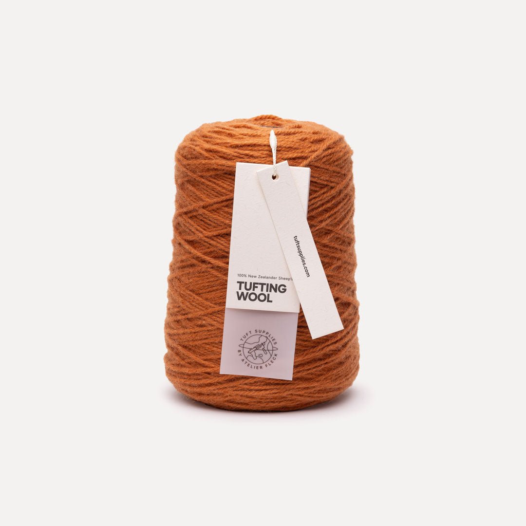 A cone of orange-brown tufting wool yarn with a white paper hangtag reading 'TUFTING WOOL' and 'tuftsupplies.com', photographed on a clean off-white background.