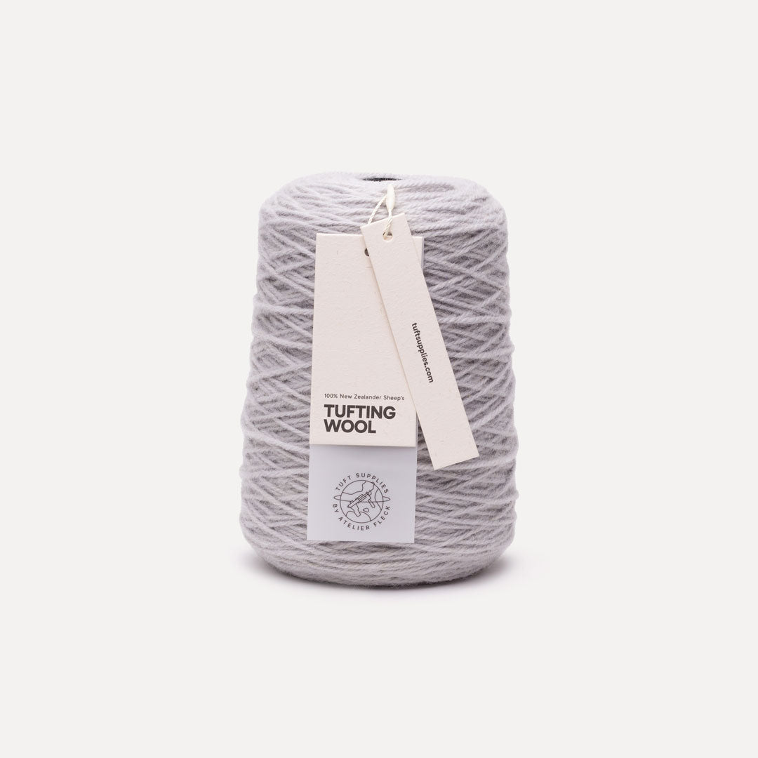 A cone of light gray tufting wool yarn with a white paper hangtag, photographed against a clean, solid off-white background.