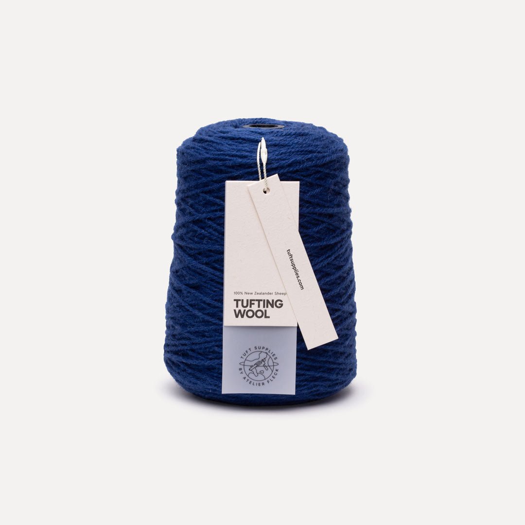 A cone of deep blue tufting wool yarn with a white paper hangtag, set against a clean off-white background.