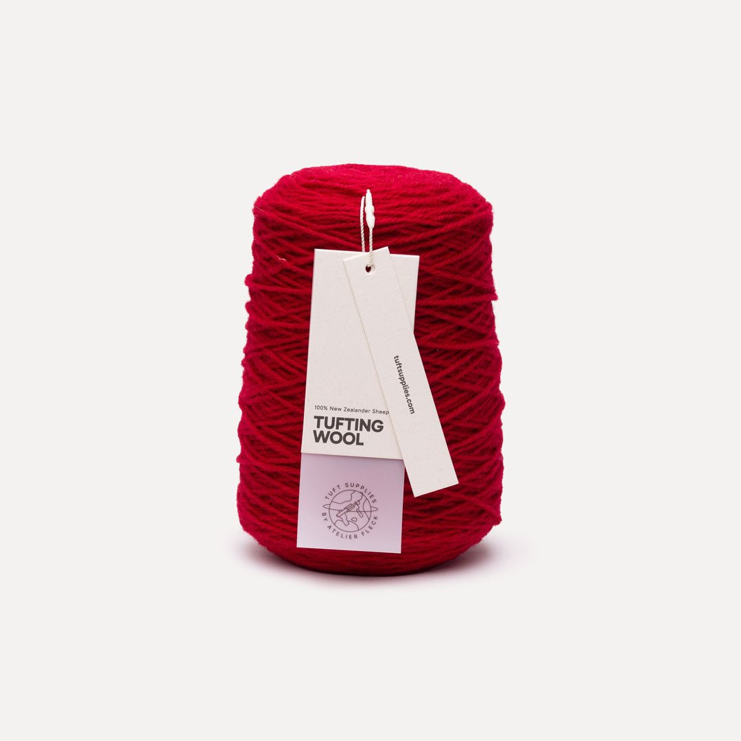 A single cone of bright red tufting wool yarn with two paper hang tags attached, photographed on a clean light gray background.