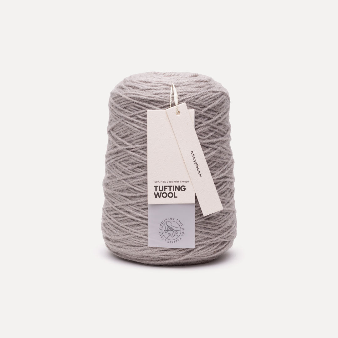A single cone of light gray tufting wool yarn with a paper hangtag, photographed on a clean, solid off-white background.