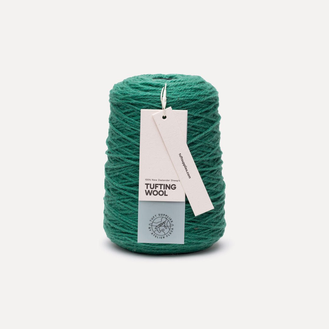A skein of teal green tufting wool yarn with a white paper hangtag, set against a clean off-white background.