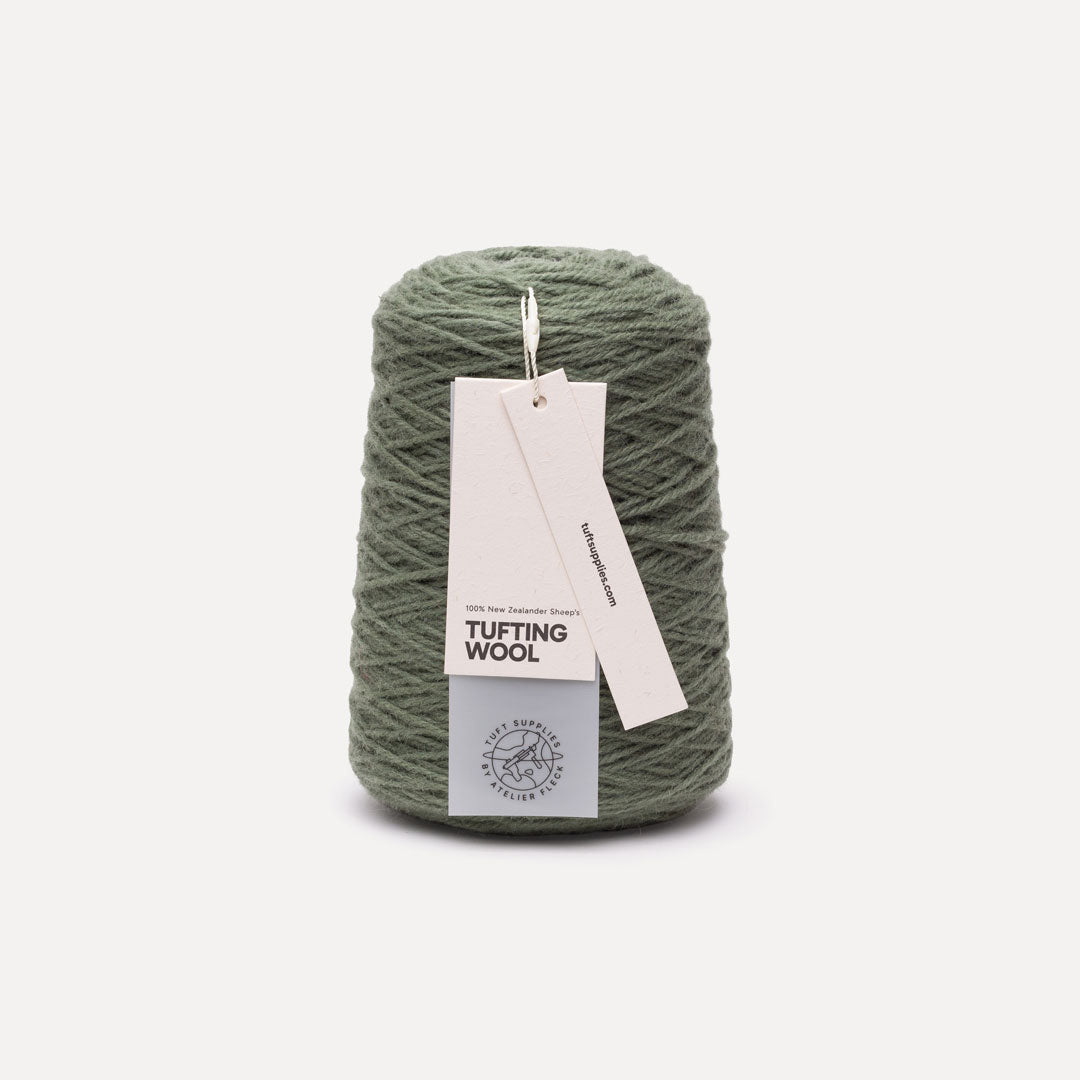 A single cone of dusty sage green tufting wool yarn with a white paper hangtag, set against a clean, light off-white background.