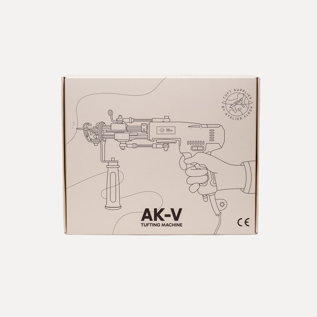 A square cardboard product box for an AK-V Tufting Machine, featuring a black line drawing of the tool and a hand on a light beige background, set against a solid off-white backdrop.