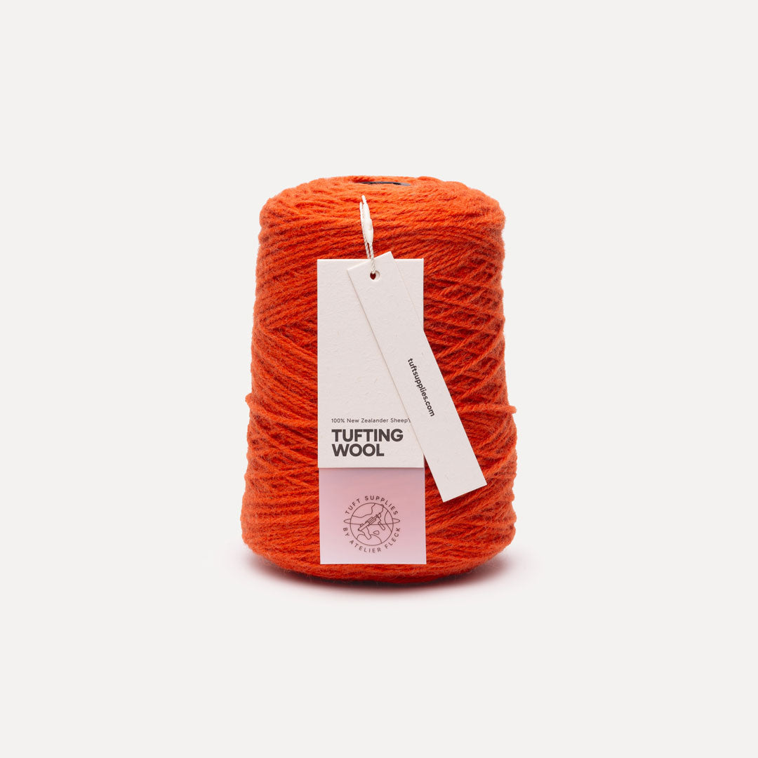 A cone of bright orange tufting wool yarn with a white paper hangtag, set against a clean, light off-white background.