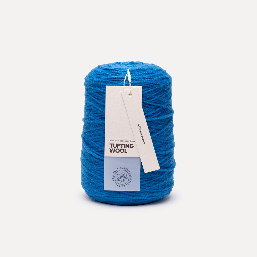 A spool of bright blue tufting wool yarn with two paper tags attached, photographed against a clean, light off-white background.